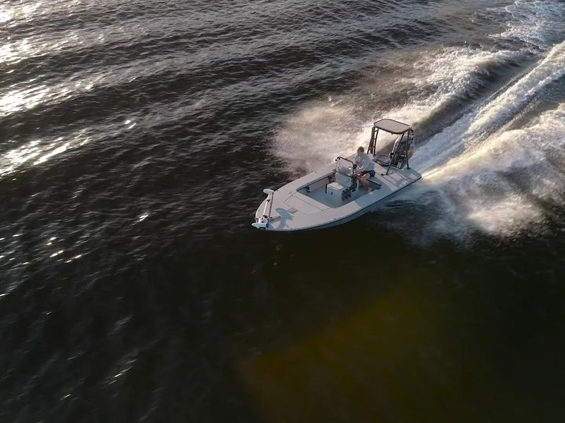 An aerial view of the small flats boat cutting across dark water with one person at the console and a white wake behind